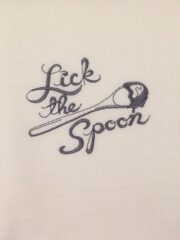 A white flour sack towel with the words "Lick The Spoon" embroidered on it.